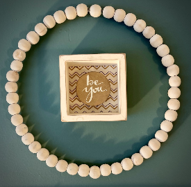 Small framed "be you" print inside a wooden-bead wreath on a blue wall
